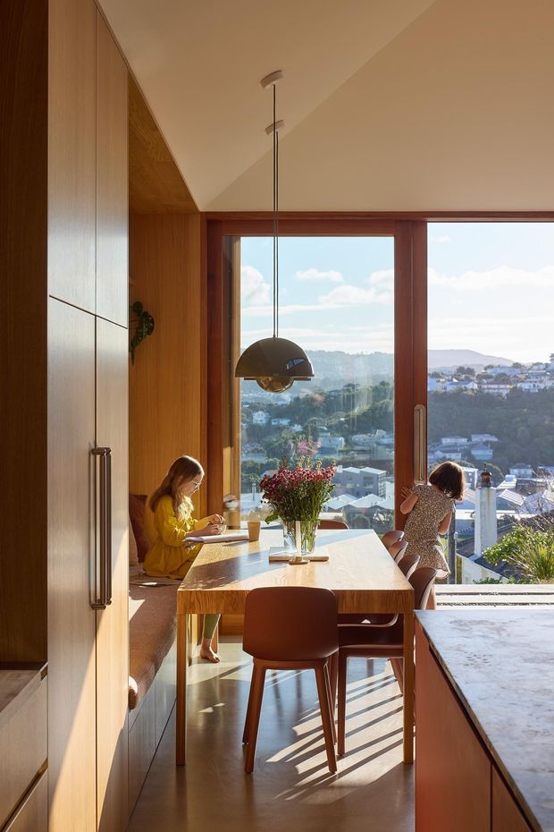Sunlit dining and kitchen area with warm timber cabinetry and wall panelling, a built-in window seat, timber dining table and chairs, and a large pendant light. Floor-to-ceiling glazing frames a hilly suburban view beyond. A stone-topped kitchen island is visible in the foreground.