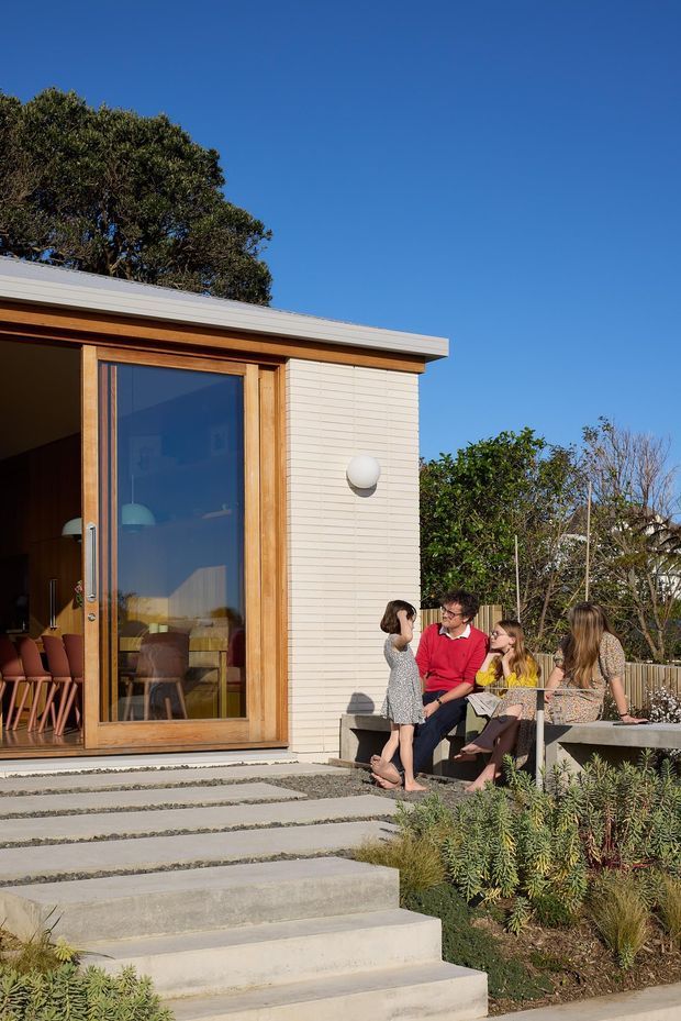 Exterior view of a contemporary single-storey home extension or pavilion opening onto a landscaped terrace. The building has pale horizontal cladding, a low-profile flat roof, warm timber-framed full-height glazing and broad concrete steps. A family is gathered on a built-in outdoor bench beside native-style planting under a clear blue sky.