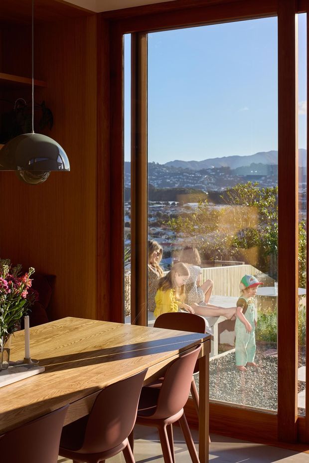 A warm, wood-lined dining area with a timber dining table, sculptural pendant light, and contemporary upholstered dining chairs. Full-height glazed doors or windows open to a sunlit outdoor area, with a suburban hillside and mountain landscape visible beyond. The image has rich timber tones, strong natural light, and a family-oriented indoor-outdoor connection.