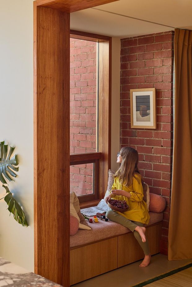A warm, intimate interior nook with a built-in timber window seat beneath a tall timber-framed window. The seat has upholstered cushions and integrated storage cabinetry, positioned against an exposed red brick wall. A person in a mustard-yellow outfit sits reading or relaxing, with curtains at the side and a framed artwork on the brick wall. The space appears connected to a bedroom, with part of a bed visible in the foreground.