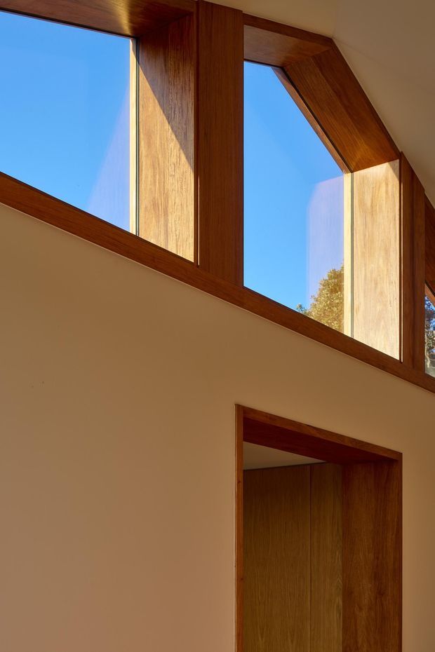 Interior architectural detail showing a tall clerestory window and doorway framed in rich dark timber. Warm sunlight falls across smooth light plaster walls, with expansive blue sky and tree foliage visible through the glazing. The image emphasizes timber joinery, geometric window framing, and natural light rather than furnishings.