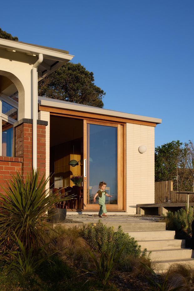 Exterior view of a contemporary residential extension with a warm timber-framed glazed door, pale horizontal cladding, exposed brickwork, and a low-pitched roof. Broad concrete steps lead through a landscaped garden to the entry, with an adjacent covered outdoor or interior transition space visible through the open doorway. The image is photographed in warm sunlight against a clear blue sky.