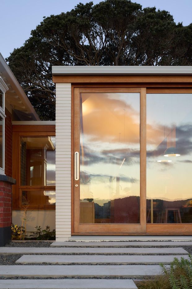 Exterior view of a modern residential extension with extensive floor-to-ceiling glazing in warm timber frames, white horizontal cladding, and a low-profile metal roof edge. Broad concrete stepping slabs set in dark gravel lead to the glazed facade. The glazing reflects a sunset sky and distant landscape, while interior pendant lights and dining furniture are faintly visible inside. Mature sculptural trees form the background.