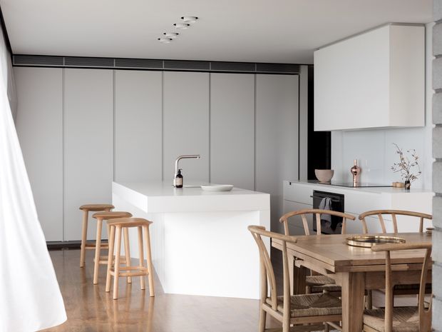 Bright, minimalist open-plan kitchen and dining area with a large white island, integrated white cabinetry, timber bar stools, and a rustic timber dining table with woven-seat chairs. Pale timber flooring, white walls and cabinetry, subtle black accents, and concealed linear ventilation create a clean, refined interior.