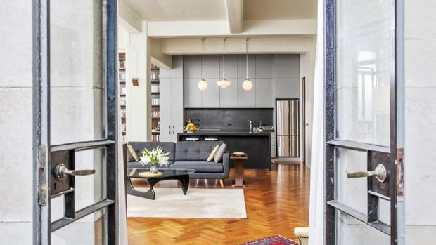 View through open glazed doors into a bright open-plan living and kitchen area. The room features a dark contemporary kitchen with stainless-steel refrigerator, charcoal cabinetry, a black leather sofa, pendant lights, exposed white ceiling beams, and warm herringbone timber flooring. A bookshelf-lined wall and colourful rug add visual interest.