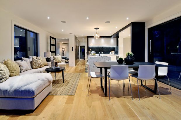 Open-plan contemporary living, dining and kitchen interior with pale timber flooring, a large sectional sofa, dining table, and a dark-framed kitchen with island. Floor-to-ceiling glazing and black-framed internal partitions create a bright yet refined evening setting.