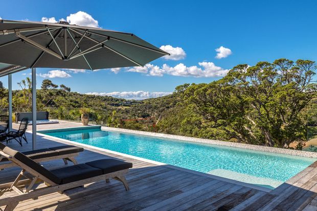 A sunlit outdoor swimming pool with turquoise water sits on a timber deck overlooking a densely wooded, rolling landscape. Large dark umbrellas shade lounge chairs beside the pool, creating a resort-style outdoor entertaining area beneath a vivid blue sky.