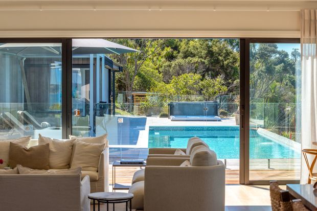 Bright, relaxed living room viewed toward a turquoise swimming pool through expansive floor-to-ceiling glass sliding doors. The room includes cream upholstered sofas and armchairs, a small dark round coffee table, warm timber flooring, and light curtains. A glazed outdoor pool terrace and landscaped greenery are visible beyond the glazing.