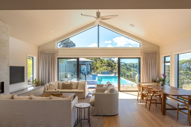 Bright, spacious open-plan living and dining area with a high pitched ceiling and dramatic triangular clerestory glazing. The room features pale upholstered sofas and armchairs, a wall-mounted television, timber flooring, a large timber dining table, and expansive glazed doors opening toward a swimming pool and leafy landscaped outlook. A ceiling fan and sheer curtains contribute to the relaxed indoor-outdoor character.