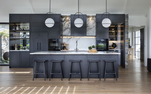 A contemporary kitchen with a large dark island and five bar stools, dark integrated cabinetry, a white marble-look splashback with veining and under-cabinet illumination, and three globe pendant lights. Timber flooring, open shelving, and adjacent living or circulation areas are visible.