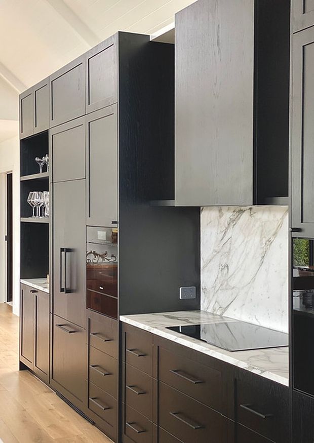 A refined modern kitchen with extensive floor-to-ceiling dark cabinetry, integrated appliances, open display shelving, and long horizontal black handles. The cabinetry combines dark timber-toned lower units with muted grey upper fronts and a contrasting black central section. A dramatic white marble-look stone benchtop and matching splashback surround the cooktop, while pale timber flooring and a white paneled ceiling brighten the space.