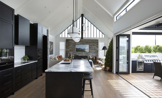 Bright open-plan kitchen and living area with a large dark island, marble-look benchtop, tall black cabinetry, pitched vaulted ceiling, clerestory windows, and a stone feature wall with a fireplace or television. The room opens through large glazed doors to an outdoor kitchen and deck.
