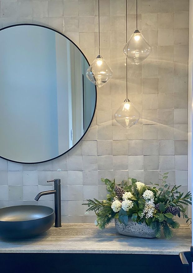 A refined bathroom vanity featuring a large round black-framed mirror, matte black vessel basin and tapware, a pale textured tiled splashback, stone-look countertop, and three suspended clear glass pendant lights. A floral arrangement adds a natural decorative element.