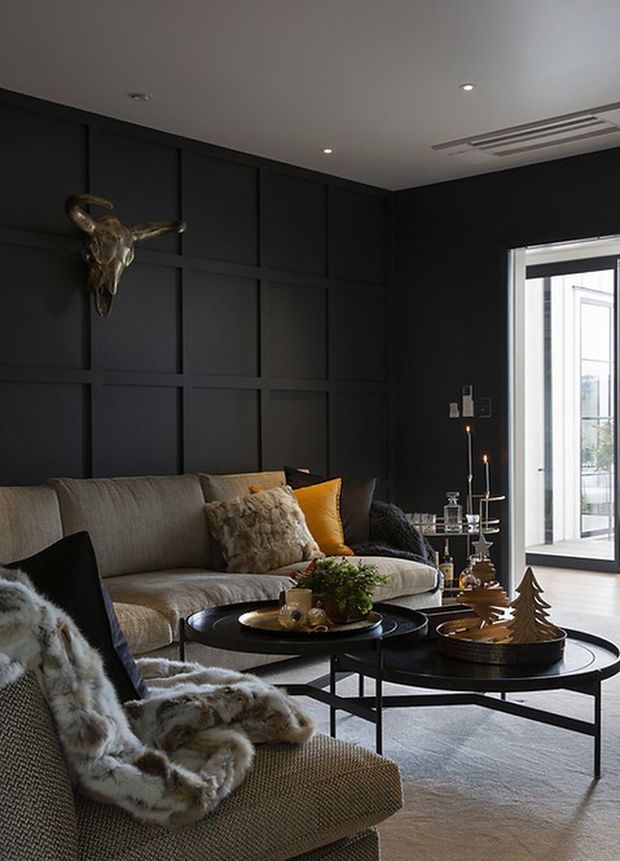 A moody, sophisticated living room featuring a large neutral sofa and lounge chair, black-painted geometric wall panelling, layered round black coffee tables, candles, sculptural decor, and dark-framed full-height glazed doors opening to a bright exterior. The room uses warm textured furnishings and metallic accents against a dark architectural backdrop.