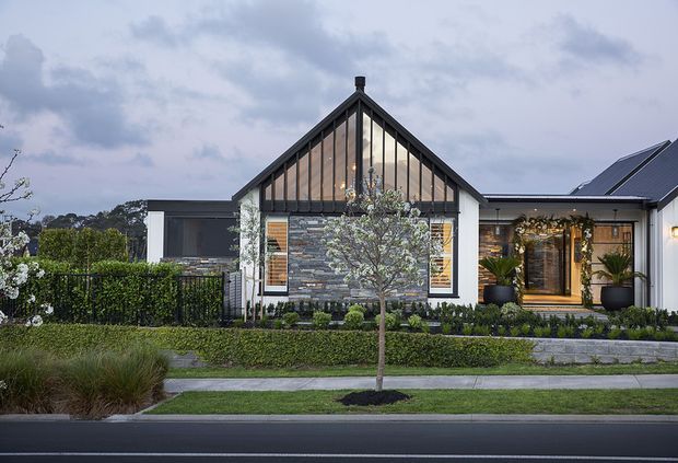 Front exterior view of a contemporary single-storey home with a prominent steep gable, dark vertical timber detailing, white walls, and a central stacked-stone feature wall. Large glazed entry doors, landscaped garden beds, hedges, a footpath, and street frontage are visible.