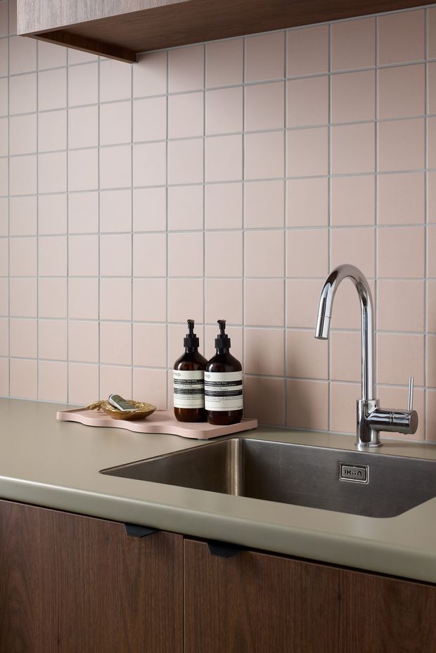 Close-up of a contemporary kitchen sink area with a deep stainless-steel undermount sink, polished chrome gooseneck tap, muted sage-green benchtop, dark timber cabinetry, and a pale blush-pink square-tile splashback. A timber overhead shelf and styled soap bottles are visible.