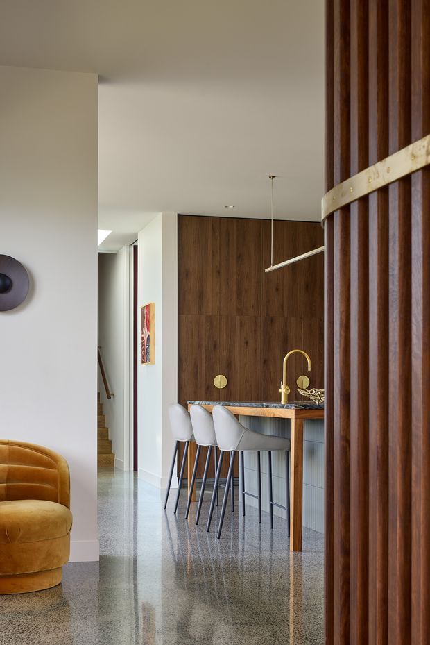 A refined open-plan interior viewed through a dark timber slatted partition. The space combines a compact kitchen or bar area with a long dark stone benchtop, integrated sink and brass tapware, timber cabinetry, and three upholstered bar stools. A caramel velvet lounge chair is partially visible in the foreground, while a stairway and hallway recede to the left. The polished speckled floor reflects the warm, contemporary interior, which is accented by brass hardware and a slim linear pendant light.