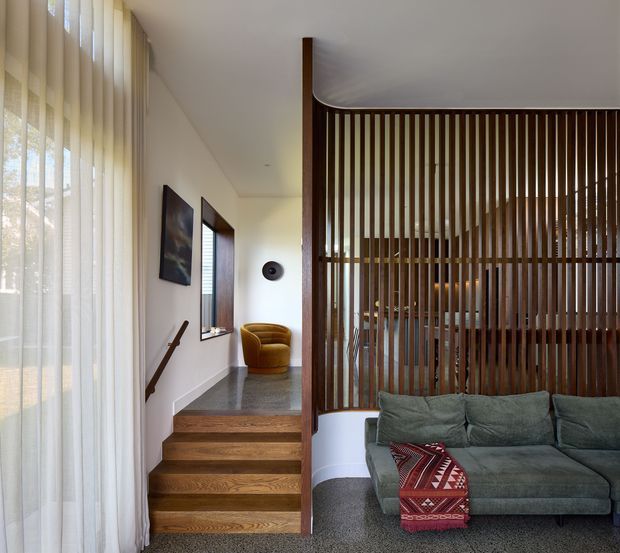 Interior view of a refined contemporary living area beside a short staircase. The room features warm timber stair treads and a dark timber vertical slatted screen dividing the living space, with a grey sectional sofa, patterned throw, sheer floor-to-ceiling curtains, white walls, and a mustard accent chair. A framed artwork and wall sconce add understated decorative detail. The slatted partition partially reveals another furnished area beyond.