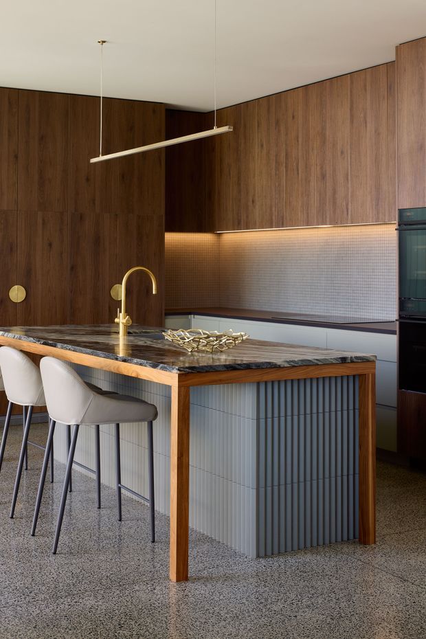 A refined contemporary kitchen centred on a large island with a dark veined stone benchtop, warm timber edging, and vertically ribbed grey cabinetry. Rich dark timber wall cabinetry and panelling extend to the ceiling, with a small square-tile splashback illuminated by concealed strip lighting. A brass-toned gooseneck tap, integrated dark appliances, white upholstered bar stools, and a slim suspended linear pendant complete the sophisticated, warm-toned interior.