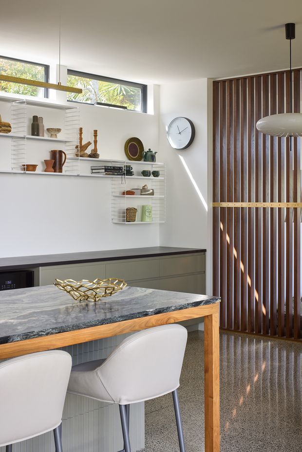 A bright, mid-century-inspired kitchen and dining area with white walls, high horizontal windows, open white shelving displaying ceramics, dark lower cabinetry, and a substantial marble island or dining table with a timber edge. White upholstered chairs sit around the island. A vertical timber slat screen separates the space, while a large sculptural pendant light and wall clock add visual interest. The polished terrazzo floor and strong natural light create a clean, refined atmosphere.