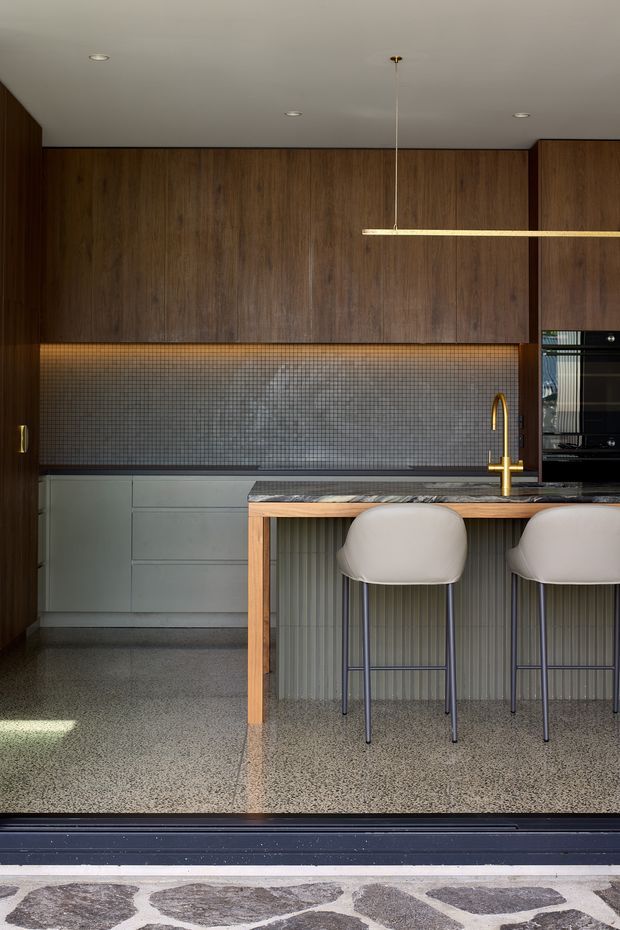 A contemporary kitchen viewed from an open threshold, featuring full-height dark timber cabinetry, a grey mosaic tile splashback with warm under-cabinet lighting, muted grey lower cabinetry, a stone island benchtop, and a timber-framed island with integrated seating for two. Two upholstered bar stools sit beneath a slim linear pendant light. The floor is a polished speckled terrazzo-like finish, and patterned exterior paving is visible beyond the doorway.
