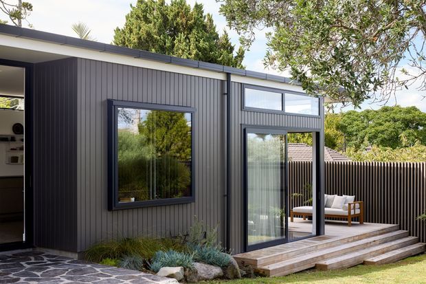 Exterior view of a contemporary single-storey house extension with dark vertical cladding, large black-framed windows and glazed doors, a low-pitched roof, and timber steps leading to a small outdoor seating area. A stone-paved path, landscaped planting, and established trees surround the addition.