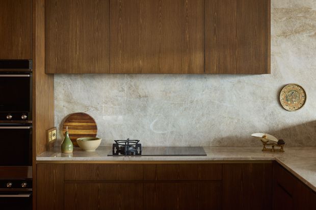 A warm, compact kitchen with dark timber cabinetry, a pale stone or plaster-look backsplash, and a stone benchtop. The cooktop is set into the counter beneath simple wood wall cabinets, with integrated dark appliances at the left and a small collection of neutral-toned decorative objects. The space has a refined, moody, mid-century-inspired atmosphere.