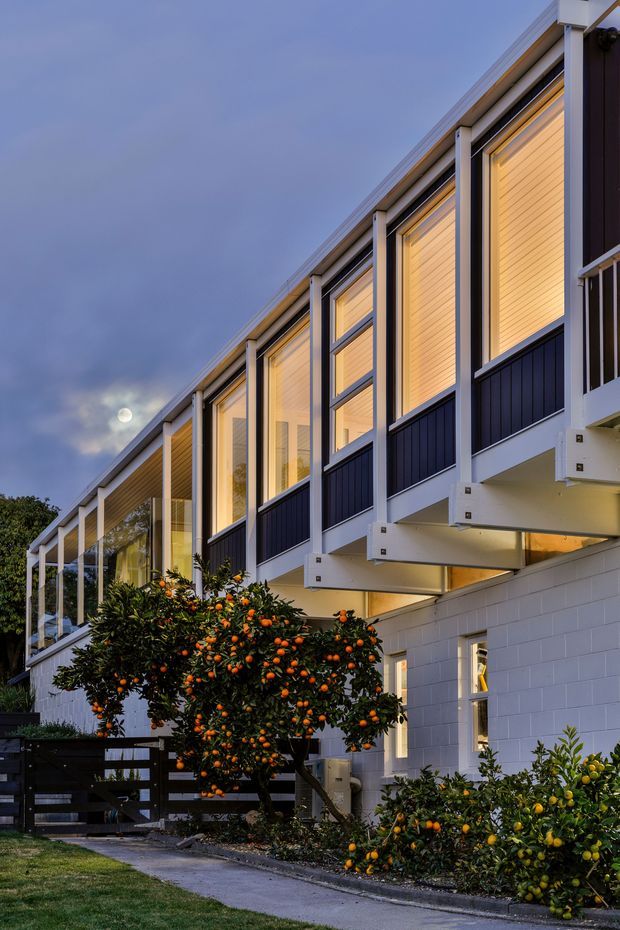 Twilight exterior view of a contemporary multi-level residential building with a long row of floor-to-ceiling glazed upper-level rooms, dark vertical cladding, white structural framing, and projecting balcony or deck platforms. The lower level has pale painted masonry walls, while citrus trees and a narrow concrete path occupy the foreground.