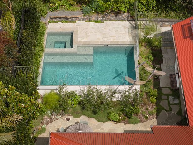 Aerial view of a landscaped residential pool area enclosed by red metal-roofed buildings. The large turquoise swimming pool has an integrated shallow step or spa zone, pale stone paving, planted garden beds, outdoor seating, and a shaded umbrella. The surrounding architecture and paths form a private courtyard-like outdoor space.