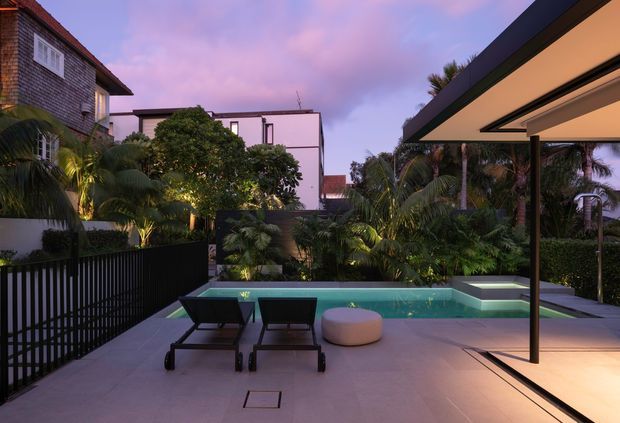 Twilight view of a landscaped residential courtyard with a compact rectangular swimming pool, paved pool deck, two sun loungers and a round ottoman. Dense tropical planting surrounds the space, with black fencing and a large cantilevered roof or covered outdoor area on the right. Neighbouring multi-storey residences are visible beyond the courtyard.