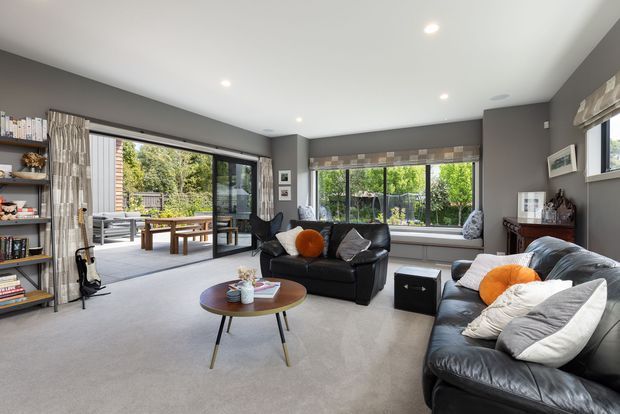 A spacious contemporary living room with dark grey walls, a white recessed ceiling with downlights, and pale carpet flooring. Black leather sofas surround a round wooden coffee table, accented by orange cushions and decorative pillows. Full-height glazing and a wide sliding glass door open to a covered outdoor entertaining area, while built-in open shelving lines the left wall.