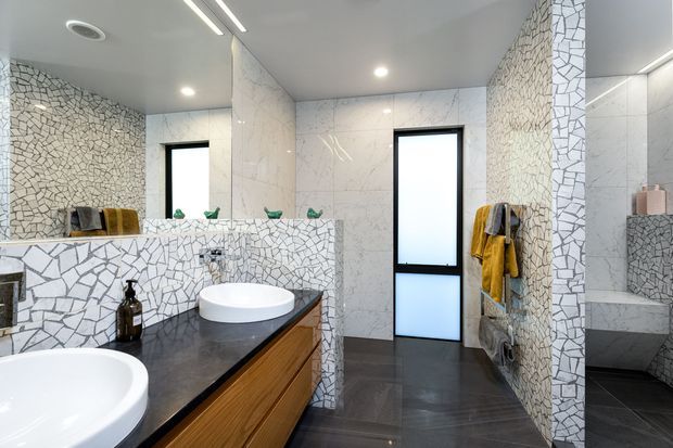 Contemporary bathroom with twin white vessel basins on a dark benchtop and warm timber vanity cabinetry. The walls feature white marble-look surfaces alongside distinctive irregular white mosaic tile cladding, with black-framed frosted windows, dark floor tiles, integrated recessed lighting, and a glass shower enclosure.