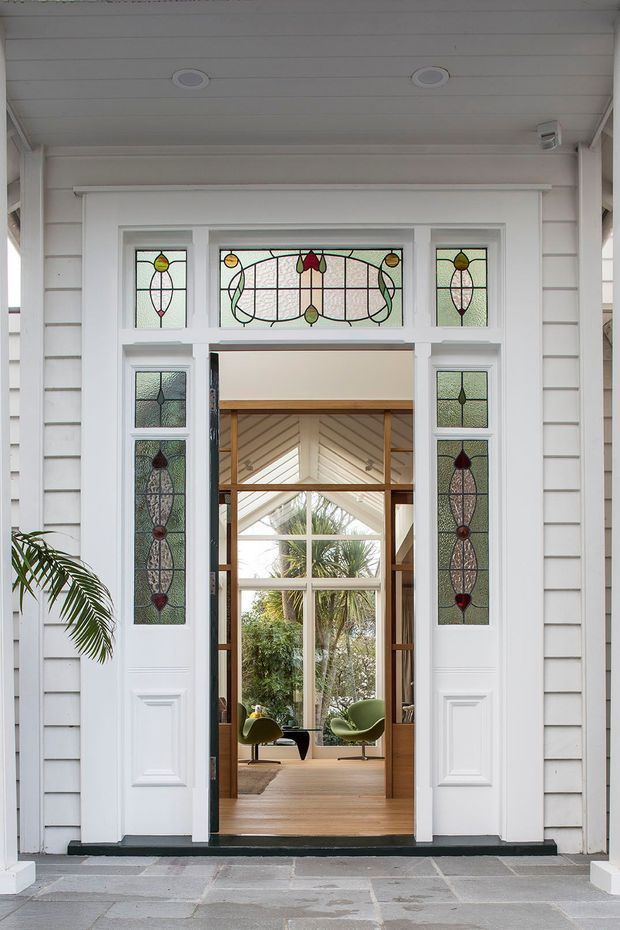 Front-facing view of a traditional residential entry porch with white weatherboard cladding, a white timber doorway, and decorative stained-glass sidelights and transom. The open door reveals a warm timber-lined interior with a glazed gable, greenery outside, and green lounge chairs. Grey stone paving forms the threshold, while recessed ceiling lights and a potted palm complete the welcoming entrance.