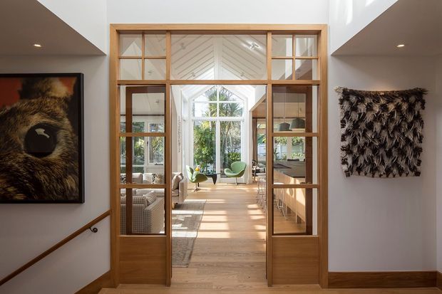 Interior view from a hallway or entry passage through glazed timber-framed double doors into a bright living area and sunroom. The space features warm timber flooring and joinery, white walls, high ceilings, abundant natural light, and a large rear window overlooking greenery. Framed artwork and a textured wall hanging flank the doorway, while a stair handrail is visible at left.