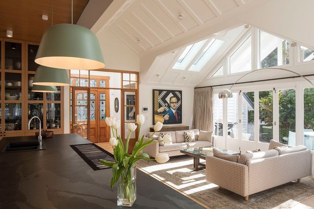 A bright open-plan kitchen and living area with a dramatic glazed sunroom extension. The kitchen features dark stone benchtops, warm timber cabinetry, integrated shelving, and oversized sage-green pendant lights. A glass-panelled timber-framed entry sits beyond the kitchen, while the living area contains pale upholstered seating, a coffee table, and a large rug. White painted timber wall and ceiling panelling, roof skylights, clerestory glazing, and abundant windows create an airy, light-filled interior.