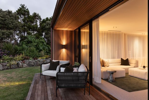 A refined indoor-outdoor living area opens onto a timber deck beside a landscaped garden. Dark-framed, floor-to-ceiling sliding glazing connects the deck to a warm, softly lit living room furnished with pale sofas, dark cushions, and a coffee table. Timber-lined soffits and vertical timber wall cladding create a cohesive contemporary retreat.