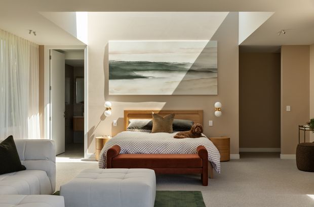 Bright, warm contemporary bedroom with a low upholstered bed, layered neutral bedding, bedside tables, wall-mounted spherical sconces, and a large abstract landscape artwork above the bed. The room opens toward a softly furnished sitting area with a white sofa and ottoman, while sheer curtains provide abundant daylight. Neutral beige and cream finishes are contrasted by warm timber and rust-toned upholstery, with geometric ceiling detailing adding architectural interest.