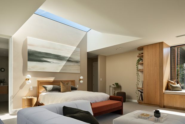 A warm, contemporary bedroom with a bed, upholstered seating, built-in timber wardrobe and shelving, and a large abstract landscape artwork. A dramatic angled ceiling opening with a skylight brings natural light into the room, while a full-height window seat and neutral finishes create a refined, inviting atmosphere.
