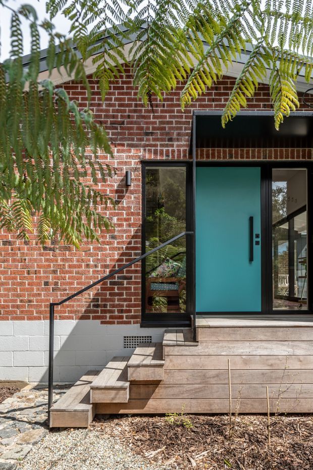 Exterior view of a contemporary brick-clad house entry. A vivid turquoise front door is set within dark-framed glazing beneath a sheltered black-clad entrance canopy. Timber decking and stepped timber stairs lead up from a gravel and mulched landscape, with a slim black handrail and leafy tree branches partially framing the facade.