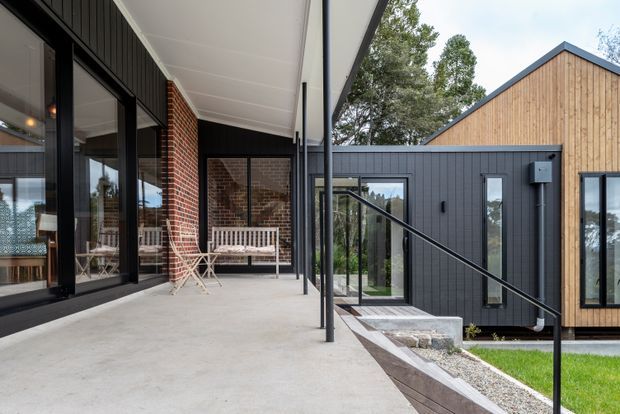 Contemporary residential exterior showing a covered concrete terrace beside a dark-clad and timber-clad house. Large black-framed glazed doors open onto the terrace, with a small outdoor dining setting and built-in bench. The building combines vertical dark cladding, warm timber weatherboards, exposed red brick, gabled roof forms, and a black metal handrail along steps leading through a landscaped courtyard.