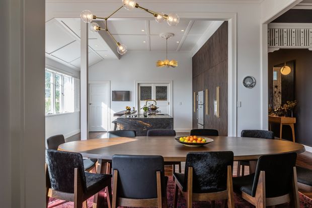 A refined open-plan dining room with a large oval timber dining table surrounded by dark upholstered dining chairs. The room features a dramatic dark timber built-in wall with integrated cabinetry and brass handles, a marble or stone kitchen island visible beyond, white decorative wall and ceiling mouldings, large windows with shutters, and sculptural contemporary pendant lighting. A darker adjoining space with a console is visible through an opening on the right.
