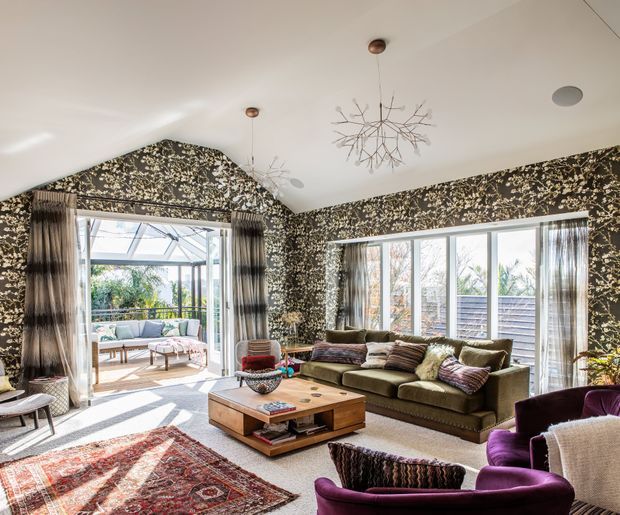 A bright, eclectic living room with a vaulted white ceiling, dramatic dark botanical wallpaper, large windows, and glass doors opening to a sunlit outdoor sitting area. The room features an olive-green sofa, deep purple lounge chairs, a timber coffee table, patterned area rugs, sheer and dark curtains, and sculptural branch-like pendant lights.