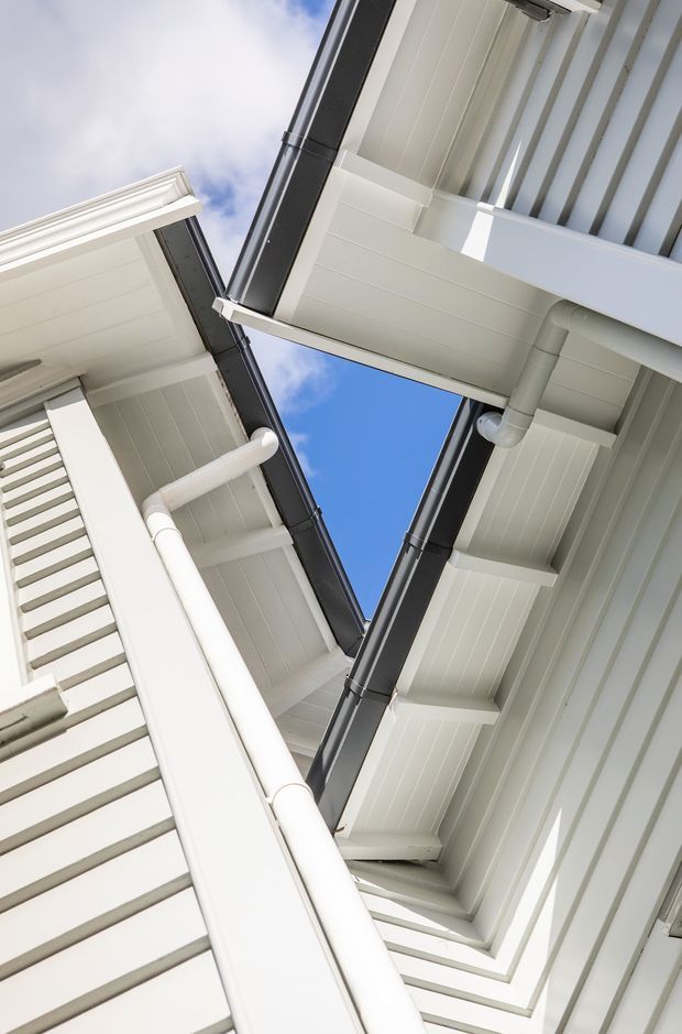 Upward-looking architectural detail showing the meeting of white weatherboard-clad building facades and projecting eaves. Dark metal gutters and downpipes contrast with the white soffits, fascia boards, exposed roof framing, and louvered exterior screening, with a narrow opening revealing blue sky.