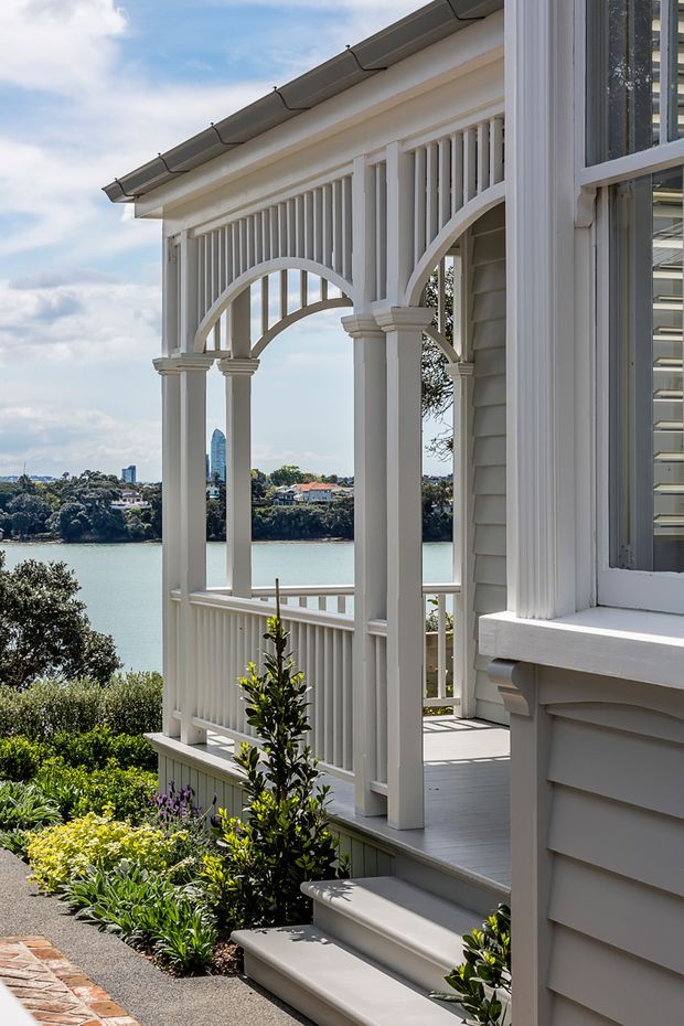 A bright exterior view of a refined white timber-clad house overlooking a broad body of water. The covered veranda features decorative arched screens, slender columns, balustrades, and a pale timber deck, with steps leading through landscaped gardens. The facade includes white weatherboards, ornate trim, and a grey pitched roof; a neighbouring shoreline and distant high-rise are visible across the water.