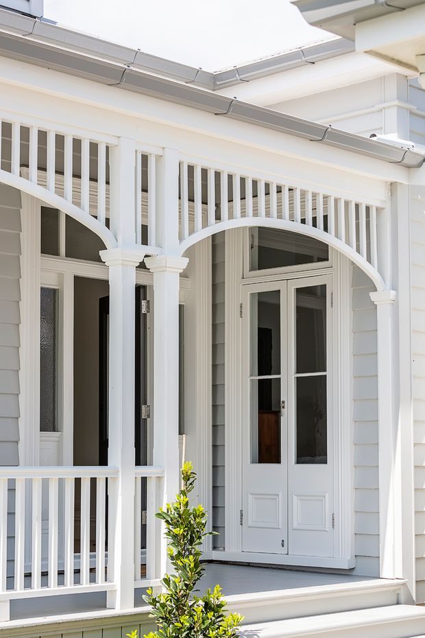 Close-up exterior view of a traditional white-painted residential porch and entry. The facade features horizontal weatherboard cladding, a covered veranda with slender square posts, decorative curved arch infill, white balustrades, a grey porch floor, and glazed French-style entry doors with transom glazing. The composition is bright and symmetrical, with a small clipped shrub in the foreground.