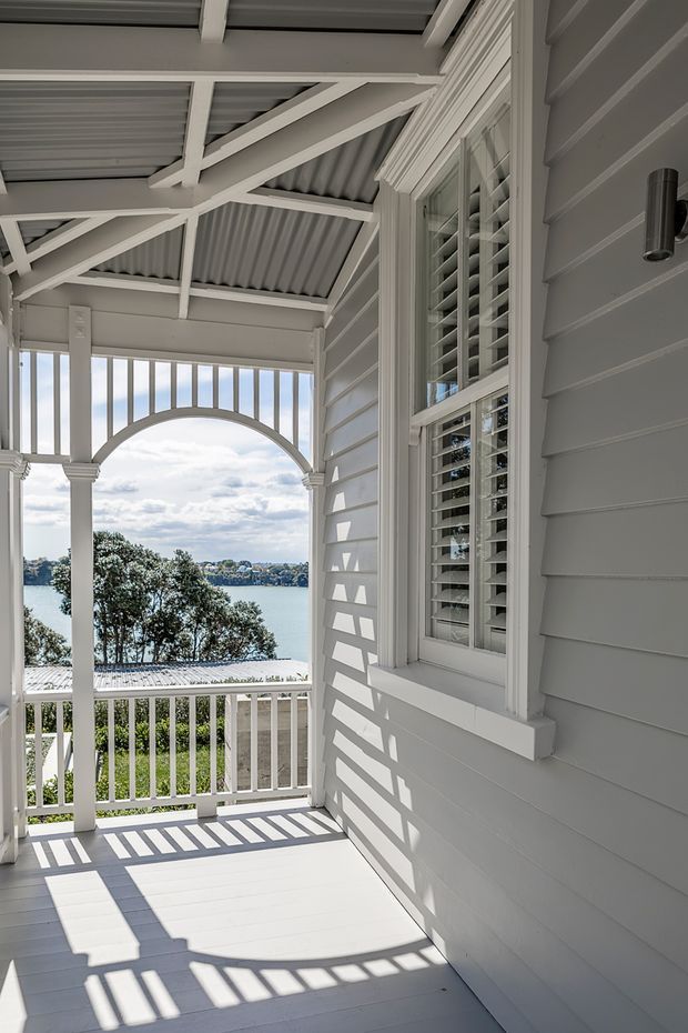 A bright covered veranda or balcony overlooking a body of water. The space features white-painted weatherboard cladding, a white timber floor, decorative posts and balustrades, exposed structural beams beneath a corrugated roof, and an arched opening with vertical spindles. A louvred/shuttered window is set into the wall, with strong sunlight casting geometric shadows across the floor and facade. Water and a distant shoreline are visible beyond the railing.