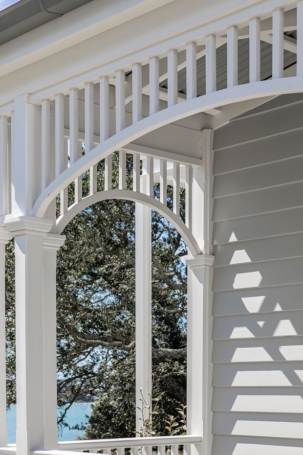 Close-up exterior architectural view of a white-painted weatherboard building with an ornate covered veranda or balcony. The facade features decorative arched timber framing, slender vertical balusters, substantial square columns with layered capitals, and a grey-white lined soffit. Bright sunlight creates geometric shadows across the wall, with trees and a glimpse of blue water visible beyond the veranda.