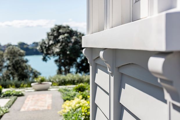 Close-up architectural exterior detail of a white-painted timber-clad building with decorative brackets and projecting trim. A landscaped garden path and water view are visible in the background, with strong sunlight creating crisp shadows across the facade.