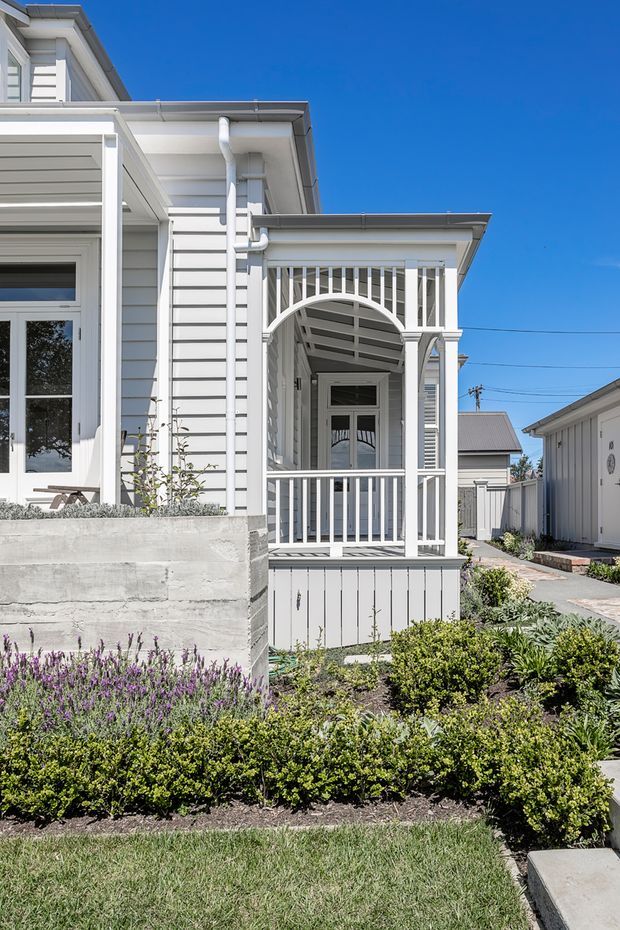Bright daytime exterior view of a white weatherboard home with a prominent covered front porch, decorative arched timber brackets, vertical balustrades, and a small landscaped front garden. The house has a traditional cottage-like facade with light grey-white cladding, white trim, pitched roof sections, and a concrete garden planter. A paved walkway leads toward the porch and adjacent outbuilding.