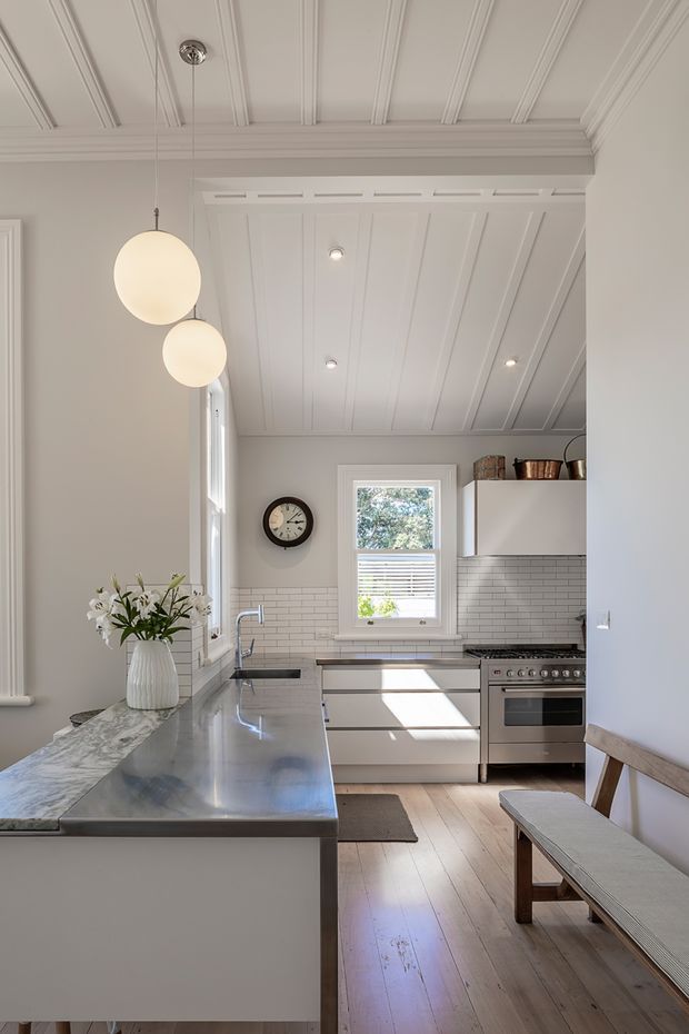 Bright, narrow traditional kitchen with white Shaker-style cabinetry, a long dark marble island, white subway-tile splashback, stainless-steel range cooker, and pale timber flooring. The room features a white panelled and beamed ceiling, detailed cornices and trim, pendant globe lights, a window above the rear benchtop, and a simple timber bench along the right wall.